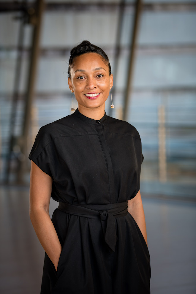 A woman in a black dress stands with hands in pockets, smiling at the camera