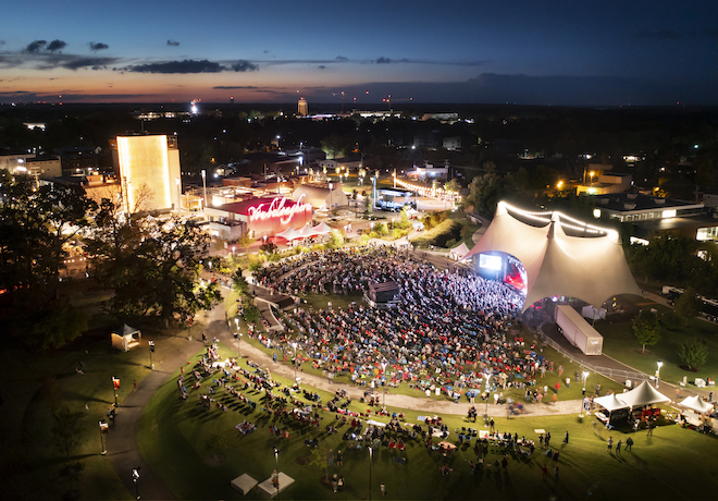 An aerial night view of a large field with a crowd of people in front of a glowing stage